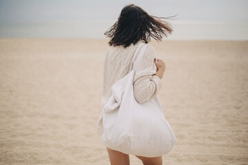 Beautiful stylish woman with windy hair and tote bag walking on sandy beach to sea, carefree...