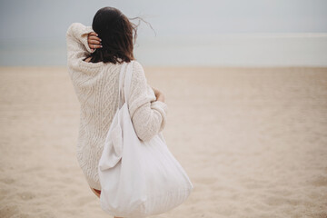 Beautiful stylish woman with windy hair and tote bag walking on sandy beach to sea, carefree...