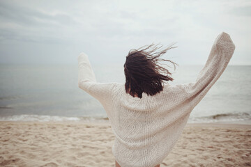 Carefree beautiful woman in knitted sweater and with windy hair running on sandy beach at cold sea, having fun. Stylish young happy female relaxing and enjoying vacation on coast. Back view