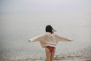 Carefree beautiful woman with windy hair in knitted sweater and bikini standing on beach at sea....