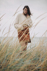 Beautiful stylish woman in knitted sweater and with windy hair posing among wild grass at sea....
