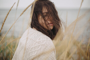Portrait of beautiful stylish woman with windy hair in knitted sweater among wild grass at sea....