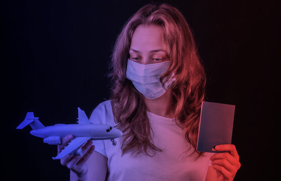 Traveling for the covid-19 pandemic. A woman in a medical mask holds a passport and an airplane figurine on a black background. Neon light