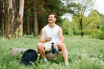 Naklejka na ściany i meble Cheerful, joyful man having a rest in nature. Smiling traveler sitting on a log in a pine forest and drinking tea from a thermos. Camping outdoors.