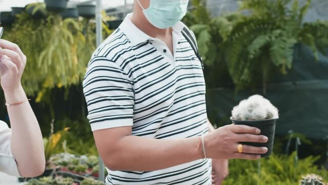 An asian man picks up a cactus in a pot at a plant shop where other people are in the background along with shelves of plants. The light is bright.