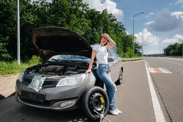 A young girl stands near a broken car with a broken wheel in the middle of the highway and is frustrated waiting for help on a hot day. Breakdown and breakdown of the car. Waiting for help.