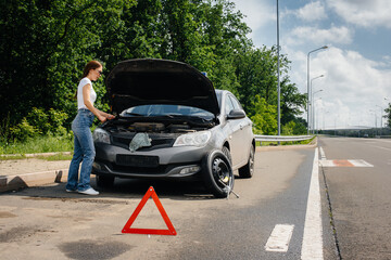 A young girl stands near a broken-down car in the middle of the highway and looks under the hood. Failure and breakdown of the car. Waiting for help.