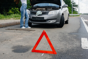 A young girl stands near a broken-down car in the middle of the highway and looks under the hood. Failure and breakdown of the car. Waiting for help.