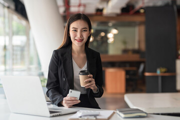 Asian businesswoman charming holding coffee mug with smartphone sitting at office. Looking at camera.
