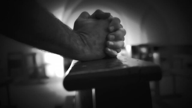 Extreme black-and-white vignette style: the clamped hands of a man praying inside a small church.
