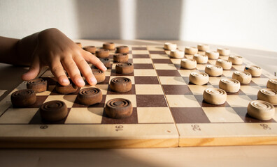 hand of a child on a wooden chessboard with white and brown checkers on a light surface of the...