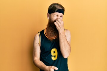 Redhead man with long beard wearing basketball uniform smelling something stinky and disgusting,...