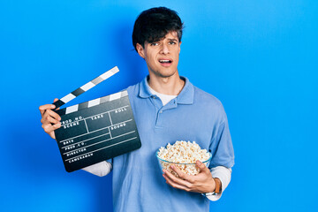 Handsome hipster young man eating popcorn holding cinema clapboard clueless and confused...