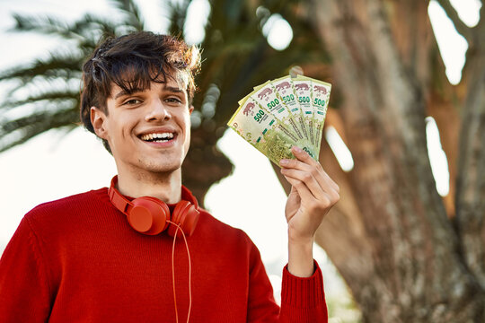 Young hispanic man using headphones holding argentinian pesos at the city