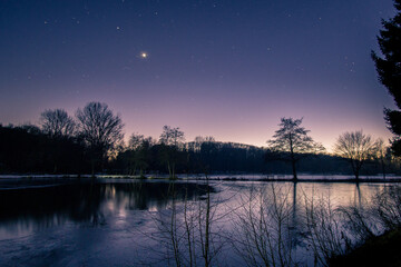 Dusk at winter evening with stars and venus on the night sky at lake landscape with silhouettes trees and reflection