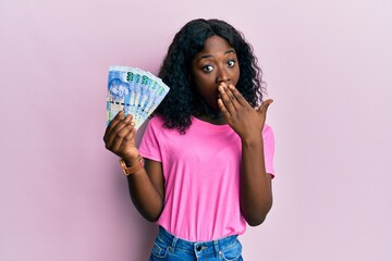 Beautiful african young woman holding south african 100 rand banknotes covering mouth with hand, shocked and afraid for mistake. surprised expression