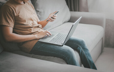 Fototapeta premium young man typing on mobile phone while sitting on a sofa at home with laptop computer. Man using his smartphone for online banking.work from home concept.