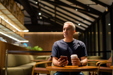 Portrait of man inside coffee shop at night thinking and using phone