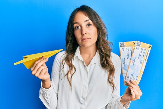 Young brunette woman holding paper airplane and boarding pass relaxed with serious expression on face. simple and natural looking at the camera.