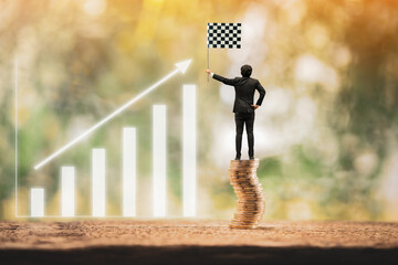 Businessman stand on the stack coin and hand hold the checkered flag a symbol of the victory for ones success and winner and arrow graph in the public park, recommend an investment goal concept.