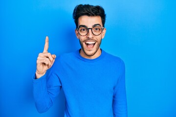 Fototapeta premium Young hispanic man wearing casual clothes and glasses pointing finger up with successful idea. exited and happy. number one.
