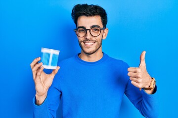 Young hispanic man holding earwax cotton remover smiling happy and positive, thumb up doing excellent and approval sign