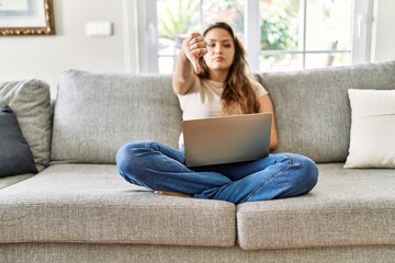 Obraz premium Beautiful young brunette woman sitting on the sofa using computer laptop at home looking unhappy and angry showing rejection and negative with thumbs down gesture. bad expression.