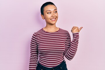 Beautiful hispanic woman with short hair wearing casual striped sweater pointing thumb up to the side smiling happy with open mouth