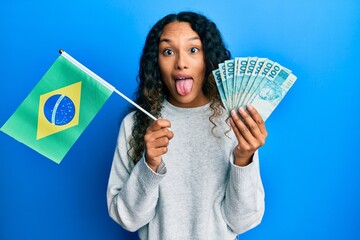 Young latin woman holding brazil flag and real banknotes sticking tongue out happy with funny...