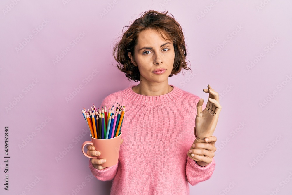 Poster Young brunette woman holding small wooden manikin hand and pencils relaxed with serious expression on face. simple and natural looking at the camera.