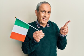 Handsome senior man with grey hair holding ireland flag smiling happy pointing with hand and finger to the side