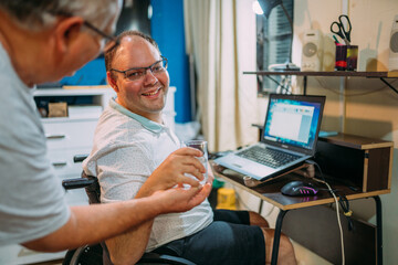 Latin man in wheelchair using laptop at home. Father taking water for his son.