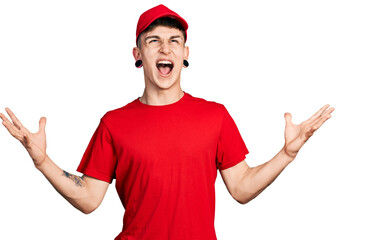 Fototapeta premium Young caucasian boy with ears dilation wearing delivery uniform and cap celebrating mad and crazy for success with arms raised and closed eyes screaming excited. winner concept