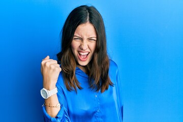 Fototapeta premium Young brunette woman wearing casual blue shirt angry and mad raising fist frustrated and furious while shouting with anger. rage and aggressive concept.