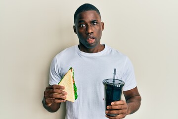 Young african american man eating sandwich and drinking soda clueless and confused expression....