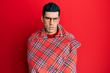 Handsome hispanic man wrapped in a red warm red blanket clueless and confused expression. doubt concept.