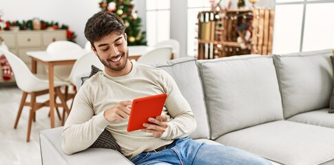 Young hispanic man smiling happy sitting on the sofa using touchpad at home.