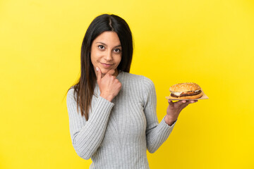 Young caucasian woman holding a burger isolated on yellow background thinking