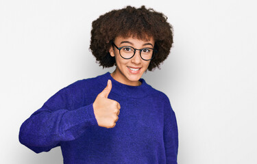 Young hispanic girl wearing casual winter sweater and glasses doing happy thumbs up gesture with...