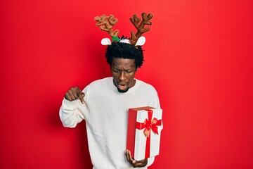 Young african american man wearing deer christmas hat holding gift pointing down with fingers showing advertisement, surprised face and open mouth