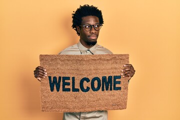 Young african american man holding welcome doormat smiling looking to the side and staring away...