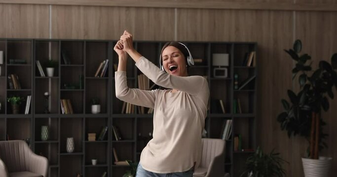 Happy carefree young female wear wireless headphones listening favourite music enjoying new hit song having fun jumping moving dancing alone in modern living room at home. Leisure and hobby concept