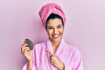 Young hispanic woman wearing shower bathrobe holding pumice stone smiling happy pointing with hand...