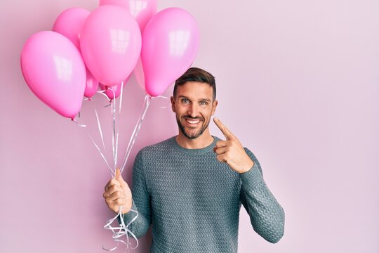Handsome man with beard holding pink balloons smiling happy pointing with hand and finger