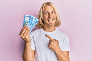 Caucasian young man with long hair holding 1000 hungarian forint banknotes smiling happy pointing with hand and finger