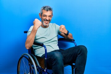 Handsome middle age man with grey hair sitting on wheelchair celebrating surprised and amazed for success with arms raised and eyes closed. winner concept.