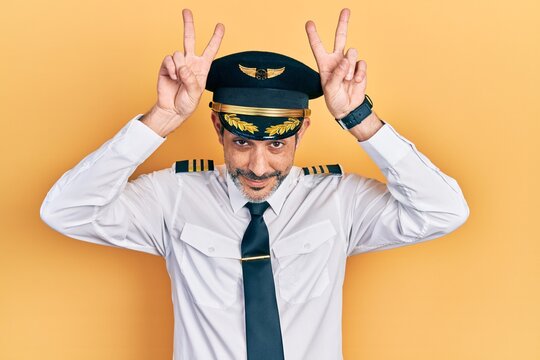 Handsome middle age man with grey hair wearing airplane pilot uniform posing funny and crazy with fingers on head as bunny ears, smiling cheerful