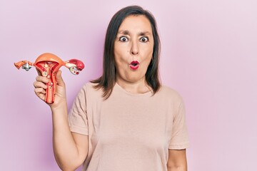 Middle age hispanic woman holding anatomical model of female genital organ scared and amazed with open mouth for surprise, disbelief face