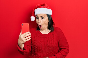 Middle age hispanic woman wearing christmas hat having video call using smartphone scared and amazed with open mouth for surprise, disbelief face