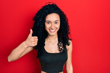 Young hispanic woman with curly hair wearing casual style with sleeveless shirt smiling happy and positive, thumb up doing excellent and approval sign